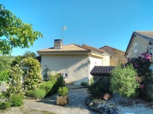 Maison de campagne avec jardin fleuri, roses roses et toit en tuiles sous un ciel bleu ensoleillé.