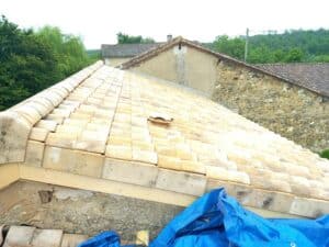 Toiture rénovée en tuiles canal beige clair sur une maison ancienne en pierre avec une bâche bleue.