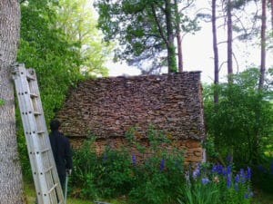 Homme devant une petite cabane traditionnelle en pierre avec toit de lauzes dans un jardin arboré.