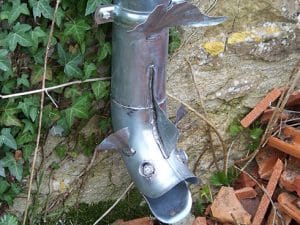 Gouttière Poisson en Métal : Décoration Jardin Originale Gouttière métallique créative en forme de tête de poisson avec nageoires sur un mur en pierre et lierre.