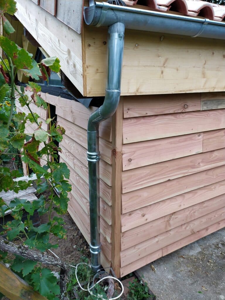 Gouttière zinc et descente pour abri de jardin en bois Gouttière en zinc installée sur le coin d'un abri de jardin en bois avec bardage horizontal et tuiles en terre cuite.