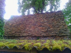 Mur traditionnel en pierre sèche sous un grand arbre avec un toit ondulé recouvert de mousse verte.
