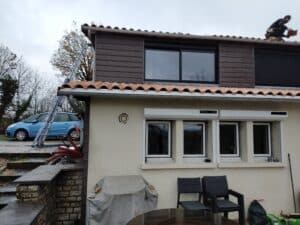 Maison avec tuiles en terre cuite, bardage sombre, fenêtres blanches et voiture bleue à côté d'une échelle de chantier.