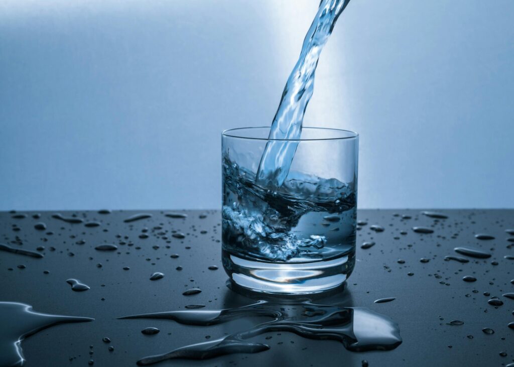 Verre d'eau rafraîchissant avec éclaboussures dynamiques Eau fraîche versée dans un verre avec éclaboussures dynamiques sur fond bleu et table mouillée.
