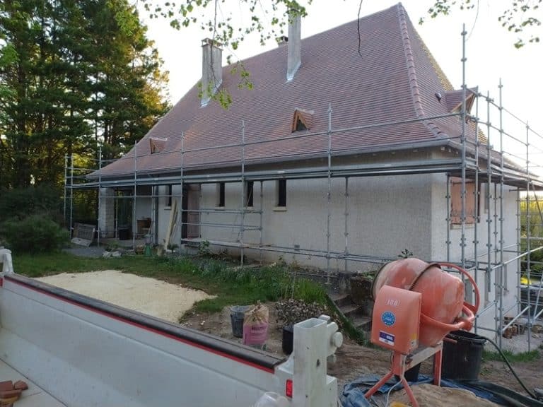 Rénovation d'une maison avec échafaudage métallique, bétonnière orange et toiture en tuiles sur un chantier extérieur.